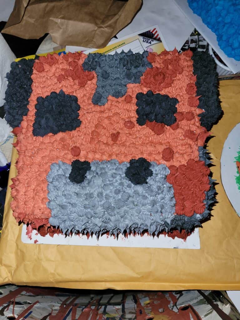 square cake decorated as a mooshroom from Minecraft, red icing covers most of cake to represent fur color, eyes, nostrils, ears are black icing, nose is grey