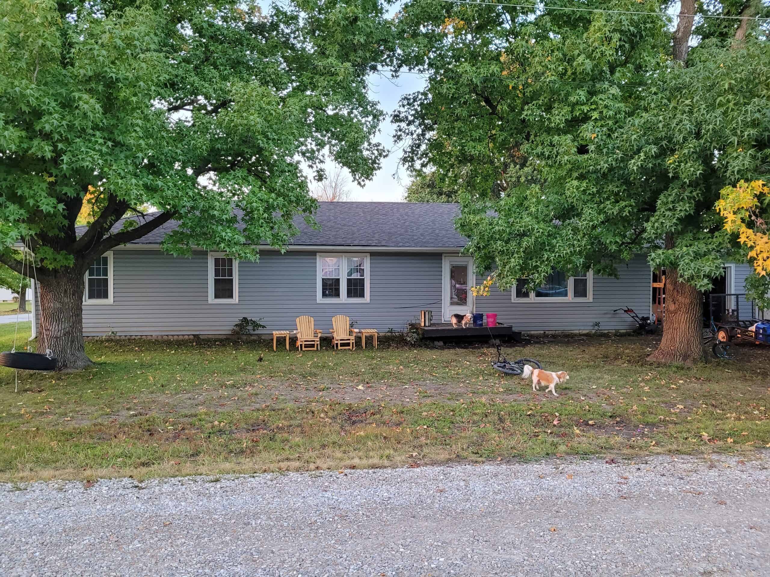a large ranch style home with blue siding and a purple front door. There are two large trees in the front yard. The house has a small deck and there is a dog running around the yard near some lawn chairs