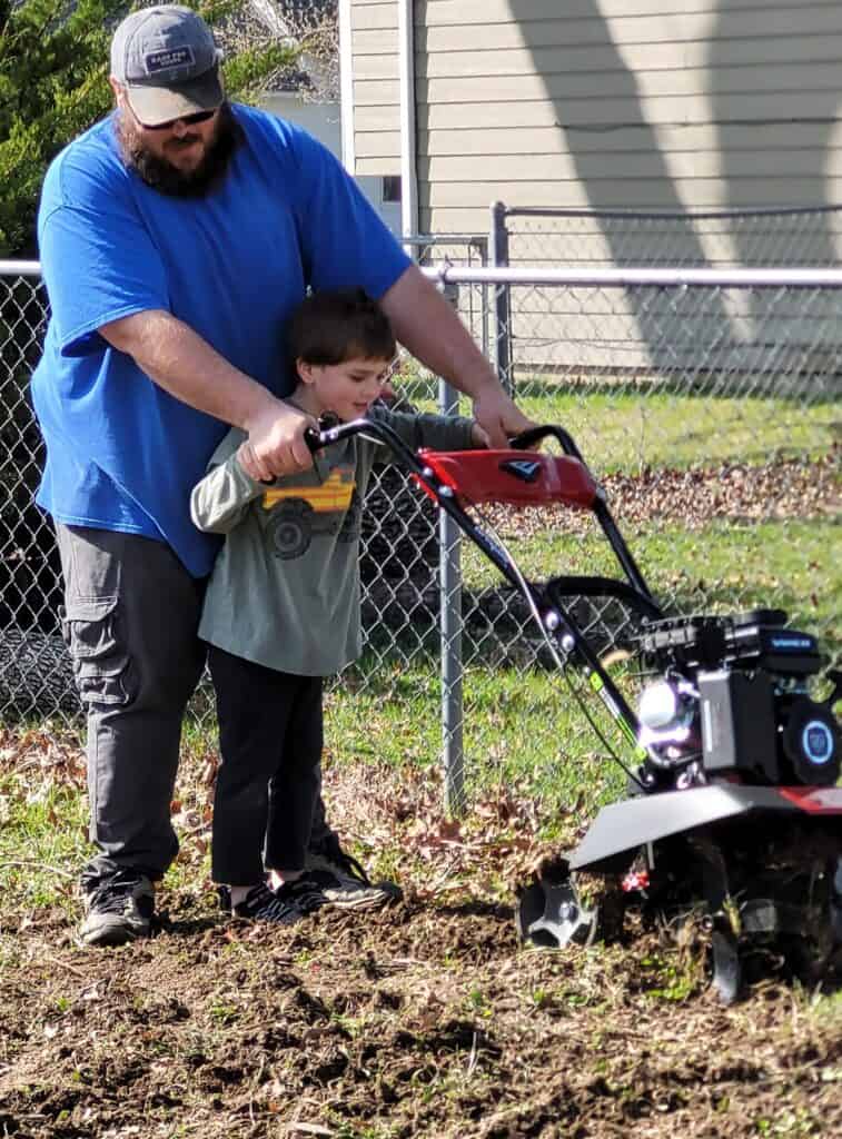 dad teaching son how to use a tiller in the vegetable garden, man is standing behind the boy behind the tiller and both are holding the handles and pushing the tiller