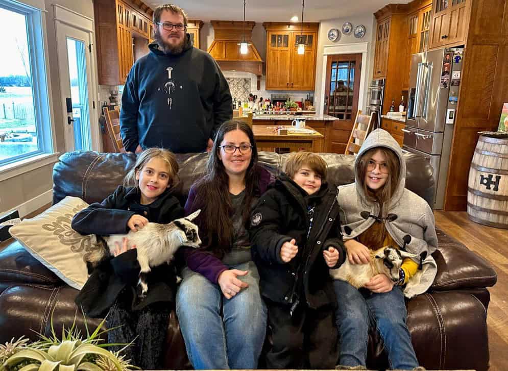 A dad standing behind a couch while his family sits on the couch holding goats, on the couch from left to right sits a girl named Abby hold a goat kid named Schweddy, then a mom and son, on the right is a girl named Kelsey, holding a goat named Jingle Bells. The goats are very small and have color patches all over that are white, brown, tan and black