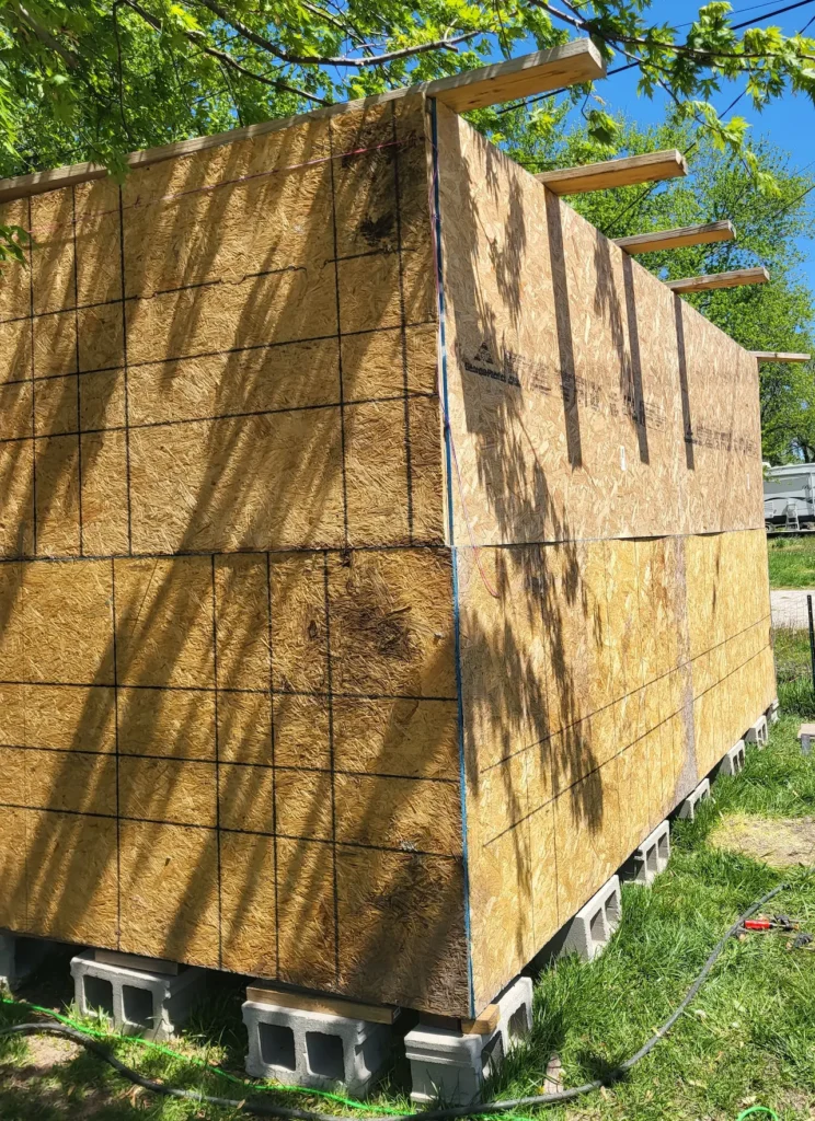 side view of goat barn showing OSB walls stacked horizontally as the wall, goat barn is anchored to several cinder blocks along the bottom of the frame