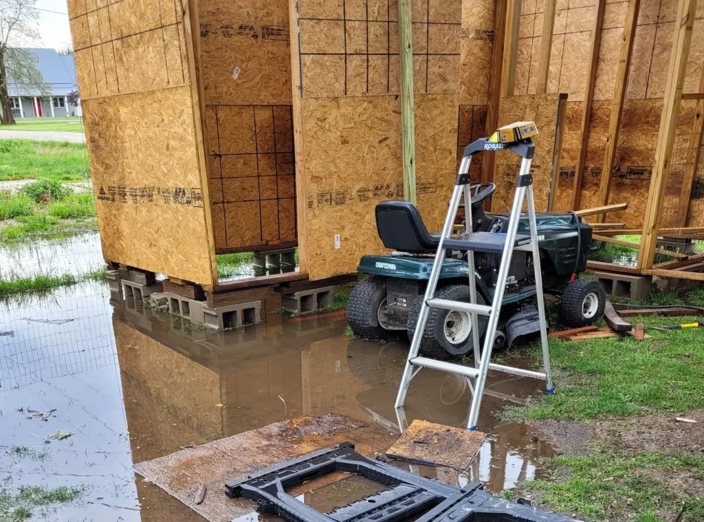 Goat barn frame and bare OSB walls sitting on cinder blocks, water pooled on ground around cinder blocks from recent rain storm. a lawn mower is parked in from of barn and a step ladder